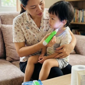 Mother using a bright green LED light earpick tool to gently clean her toddler's ear indoors.