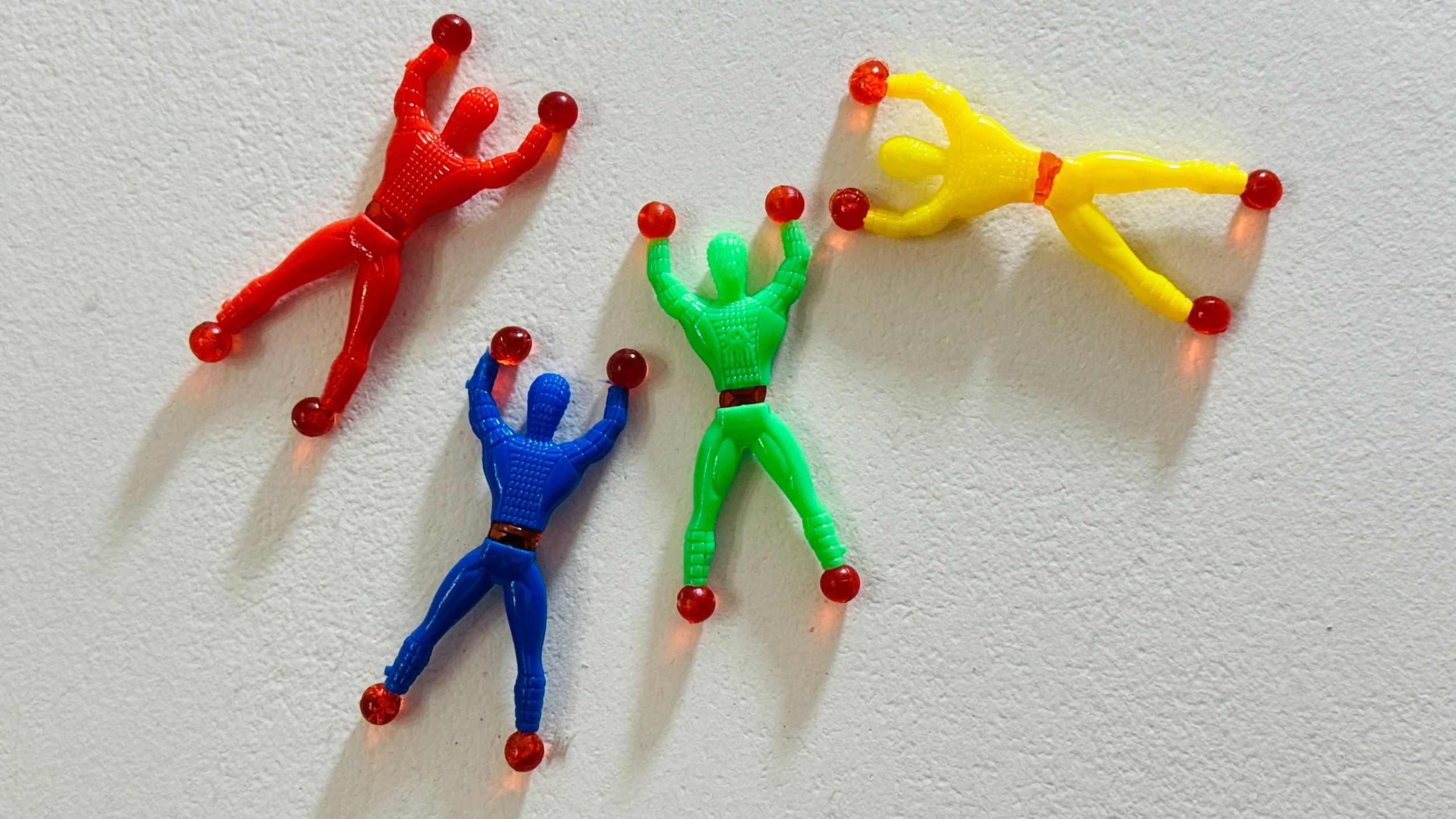 Four colorful Sticky Wall Crawler Spider-Man toys in red, blue, green, and yellow tumbling down a white textured surface.